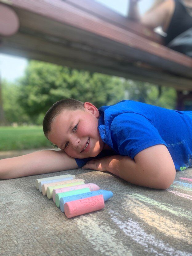 A boy in a blue shirt poses with his sidewalk chalk drawing at Arc of the Ozarks summer camp.